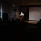A distinguished man in his 70s with white hair stands at a lectern while giving a talk in a darkened amphitheater.