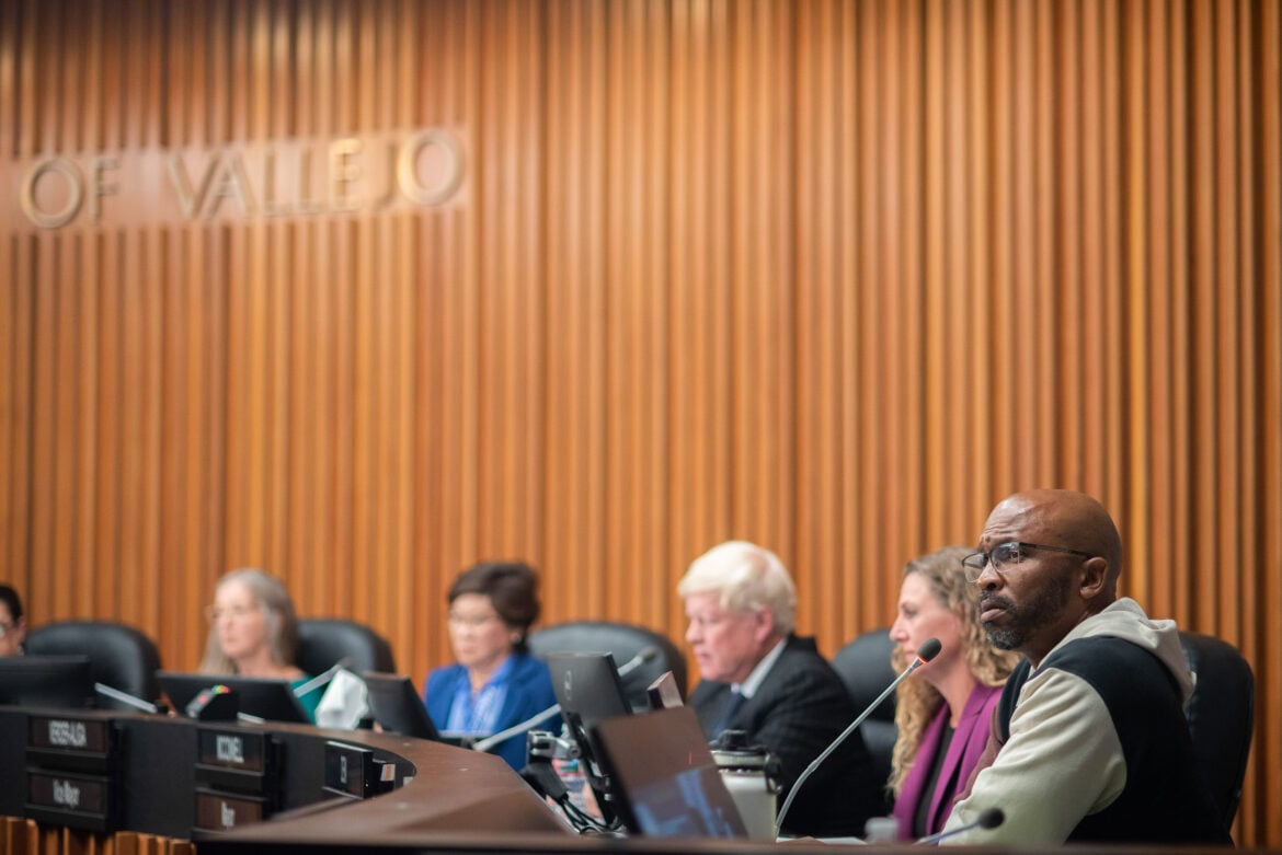 Then-Councilmember Hakeem Brown, who is a Black man in his mid-40s, listens to a speaker out of frame from the dais of the Vallejo City Council chambers. Brown, who is in sharp focus, is wearing glasses and a hoodie, and sports a closely-cropped beard. His colleagues, who are out of focus, are seen behind him. A "City of Vallejo" sign is partially visible on the wall.