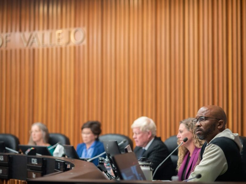 Then-Councilmember Hakeem Brown, who is a Black man in his mid-40s, listens to a speaker out of frame from the dais of the Vallejo City Council chambers. Brown, who is in sharp focus, is wearing glasses and a hoodie, and sports a closely-cropped beard. His colleagues, who are out of focus, are seen behind him. A "City of Vallejo" sign is partially visible on the wall.