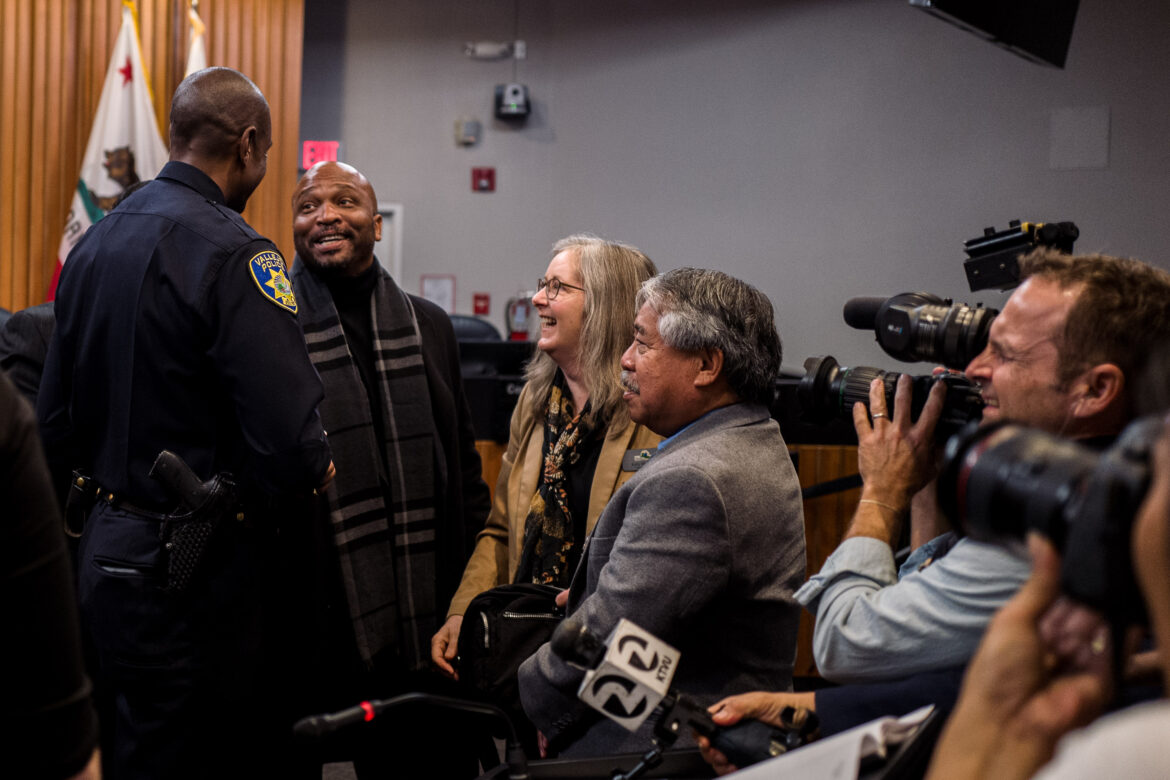 A Black councilmember in his 40s smiles excitedly while speaking with the City of Vallejo's first Black police chief inside a city council chambers while surrounded by other councilmembers and members of the news media.
