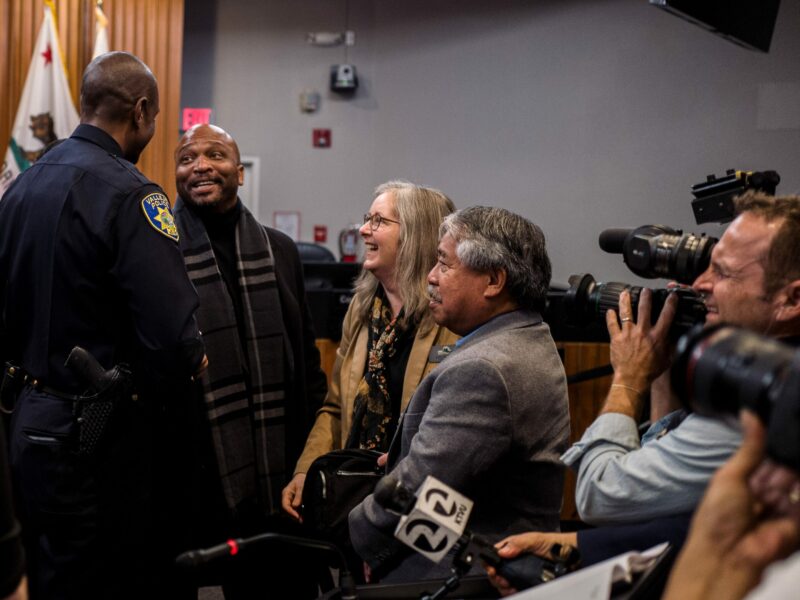 A Black councilmember in his 40s smiles excitedly while speaking with the City of Vallejo's first Black police chief inside a city council chambers while surrounded by other councilmembers and members of the news media.