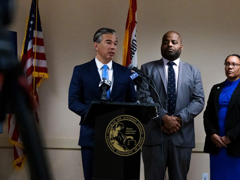 California Attorney General Rob Bonta speaks at a lectern featuring the seal of his office. The flags of California and the United States of America hang on poles behind him.