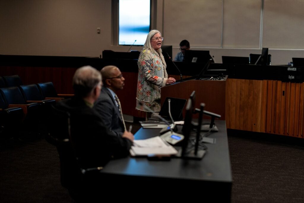 A white woman in her 50s with gray hair and wearing a floral print dress speaks from a public comment lectern at a city council meeting.