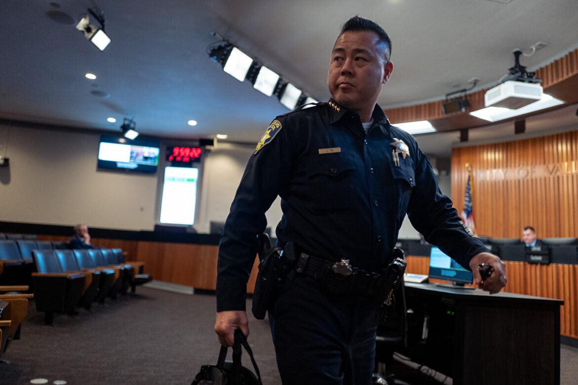 Vallejo Police Chief Jason Ta leaves a Vallejo City Council meeting, bag of documents in hand. The images catches him mid-stride. He is an Asian American man in his 50s in a dark blue police chief's uniform. The first several rows of seats are nearly empty of audience members.