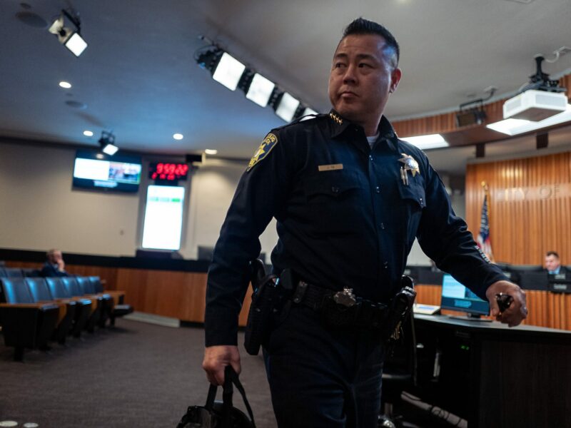 Vallejo Police Chief Jason Ta leaves a Vallejo City Council meeting, bag of documents in hand. The images catches him mid-stride. He is an Asian American man in his 50s in a dark blue police chief's uniform. The first several rows of seats are nearly empty of audience members.