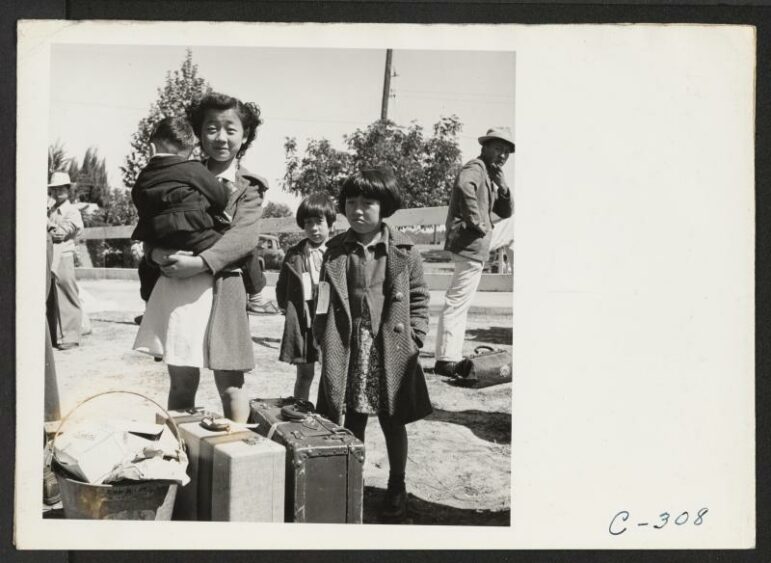 A black and white photograph of a young Japanese American girl holding a baby while standing next to her younger siblings.