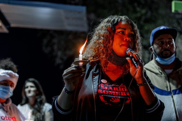 A young Black woman wearing a "Justice for Mario Romero" shirt speaks into a microphone at a nighttime gathering with other families impacted by police violence. She is holding a single candle that burns with a tall flame, lighting her face in a warm glow.