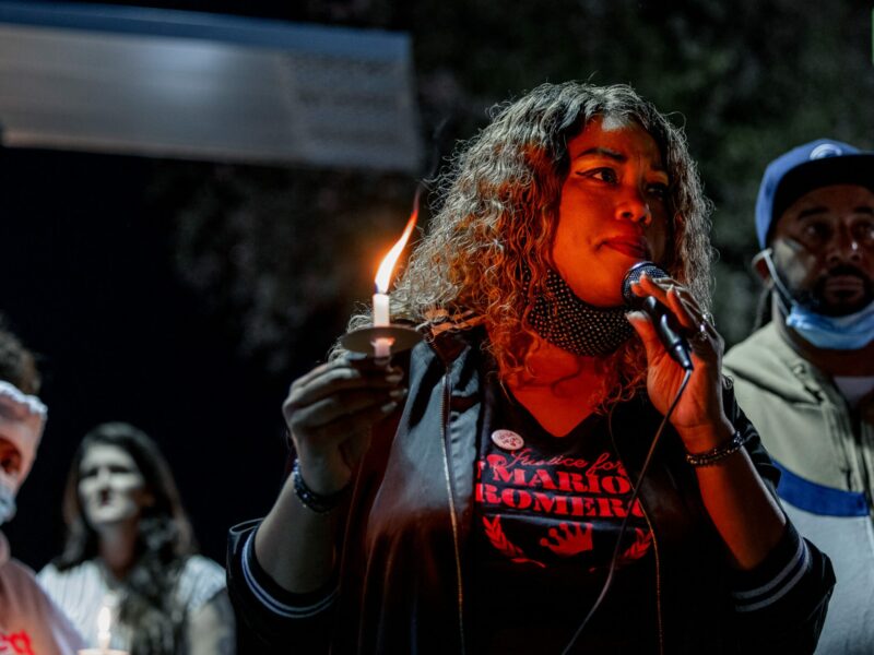 A young Black woman wearing a "Justice for Mario Romero" shirt speaks into a microphone at a nighttime gathering with other families impacted by police violence. She is holding a single candle that burns with a tall flame, lighting her face in a warm glow.
