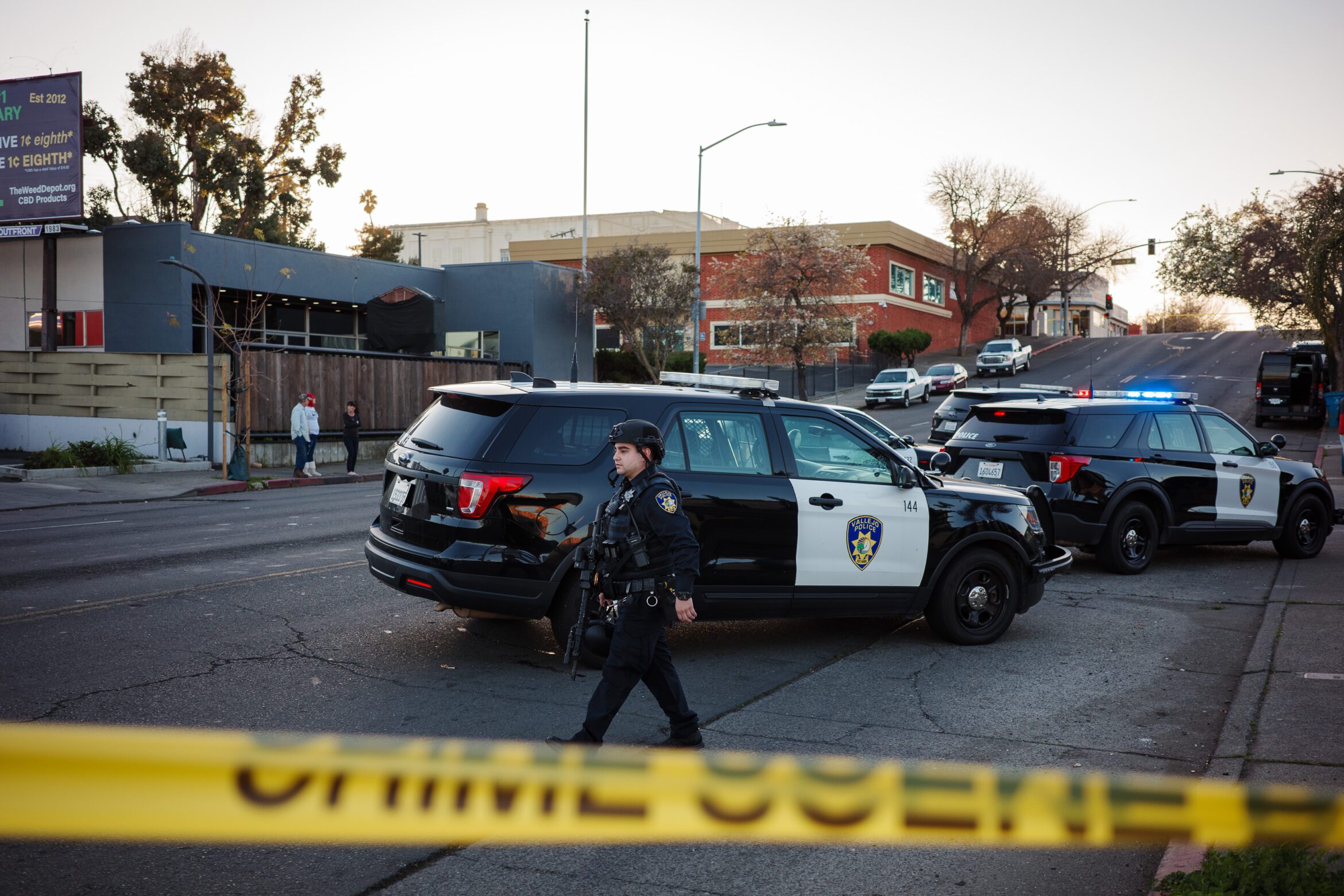 A Vallejo police officer wearing a helmet and carrying a rifle walks past police SUVs as the sun sets in Vallejo, California. Crime scene tape can be seen in the foreground.
