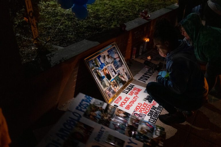 Two people bend down to look at a collage of photographs at a nighttime memorial for Ronell Foster, who was killed by Vallejo police Ofc. Ryan McMahon on Feb. 13, 2018. The scene is dimly lit save a cell phone light that illuminates a sign which reads, "Stop Police Brutality, "11.18.84," "Say His Name," "Hold Police Accountable," and "Save Our Loved Ones."