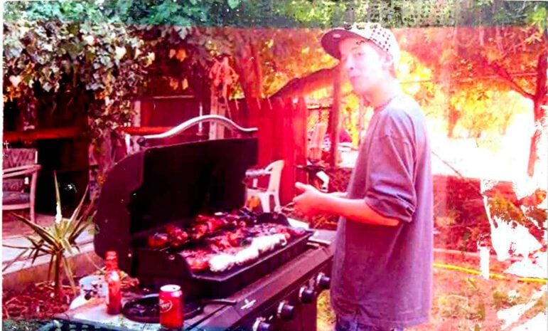 Jared Huey, a skinny teenage boy wearing a baseball cap and a longsleeve shirt with the sleeves rolled up barbecues in a backyard on a sunny day. An open Dr. Pepper soda can be seen within arm's reach. The photograph appears to have been taken with a broken camera that let light leak in, giving the image a nostalgic, multi-color effect.