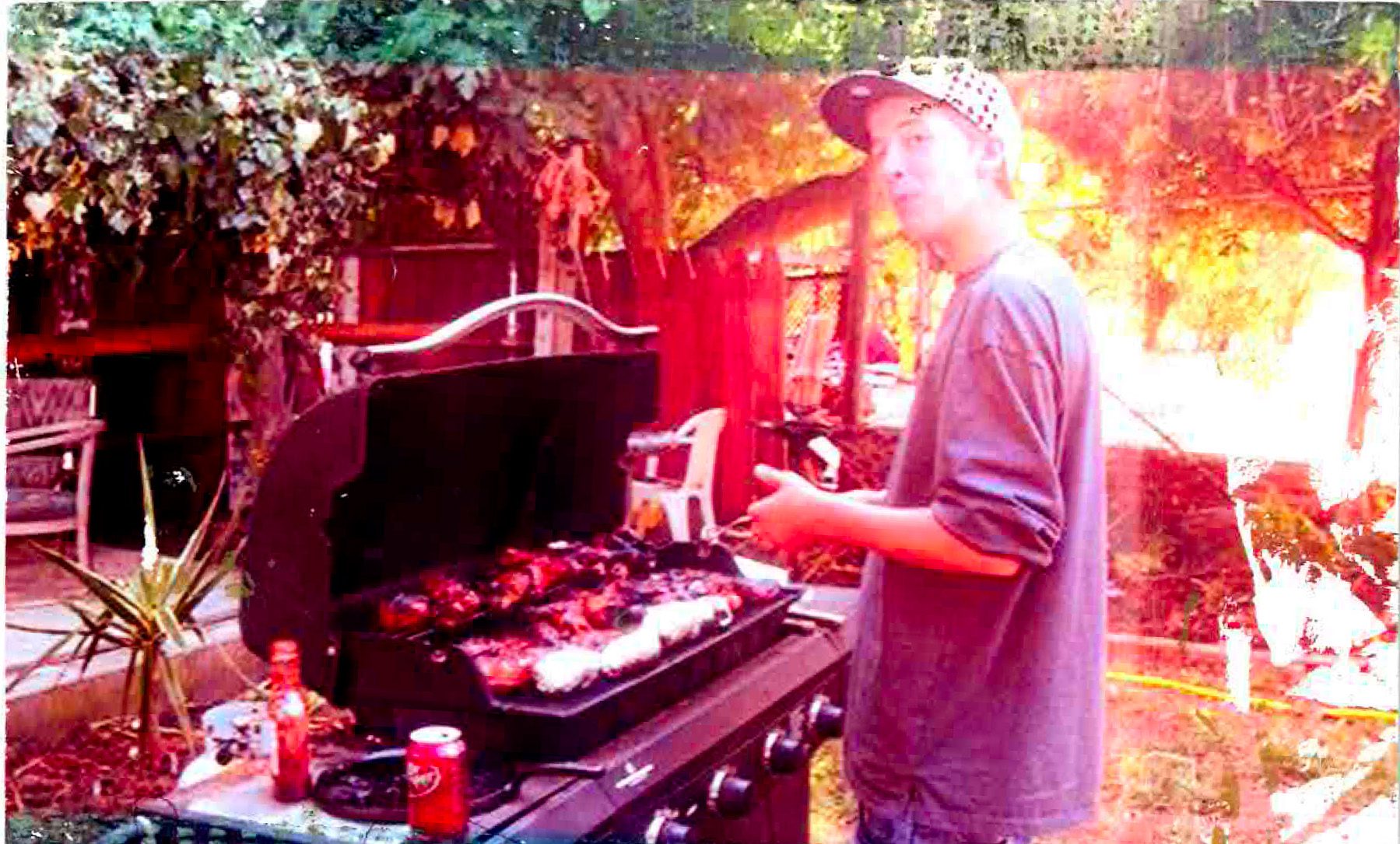 Jared Huey, a skinny teenage boy wearing a baseball cap and a longsleeve shirt with the sleeves rolled up barbecues in a backyard on a sunny day. An open Dr. Pepper soda can be seen within arm's reach. The photograph appears to have been taken with a broken camera that let light leak in, giving the image a nostalgic, multi-color effect.