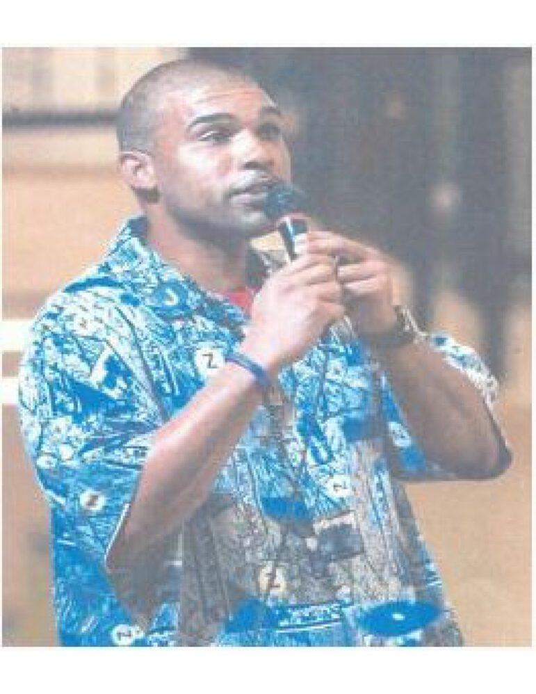 34-year-old Guy Jarreau, a Black man, is seen speaking into a microphone at an event indoors. He is wearing a patterned, bright blue shirt.
