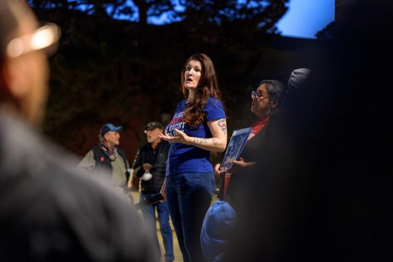 A brunette woman in her 40s wearing a blue t-shirt that reads, "No justice, no peace, no racist police" speaks to a crowd outside in the early evening.