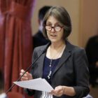 California State Sen. Nancy Skinner speaks at a podium inside California's Capitol while holding a sheaf of paper and wearing glasses.