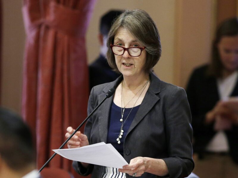 California State Sen. Nancy Skinner speaks at a podium inside California's Capitol while holding a sheaf of paper and wearing glasses.
