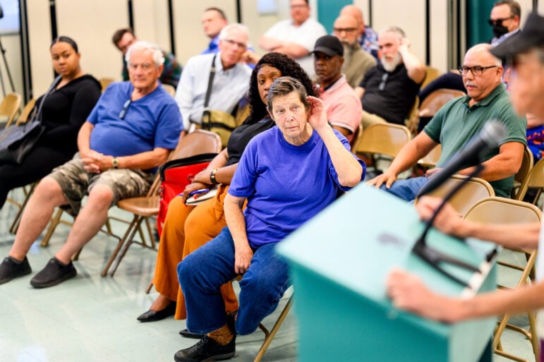 A woman with gray hair wearing a purple shirt cups her ear among a diverse crowd during a town hall in an elementary school.