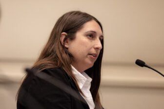 An attorney in her 30s with brown hair wearing a white shirt and dark blazer speaks into a microphone in court.