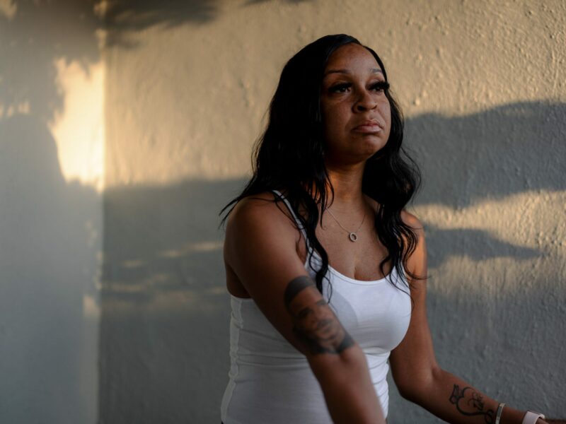 A Black woman in her early 40s wearing a white shirt poses for a portrait on her porch at sunset. An uncannily-accurate tattoo of her twin brother Charles, who was killed by Vallejo police in 2007, when they were 26, adorns her right arm.