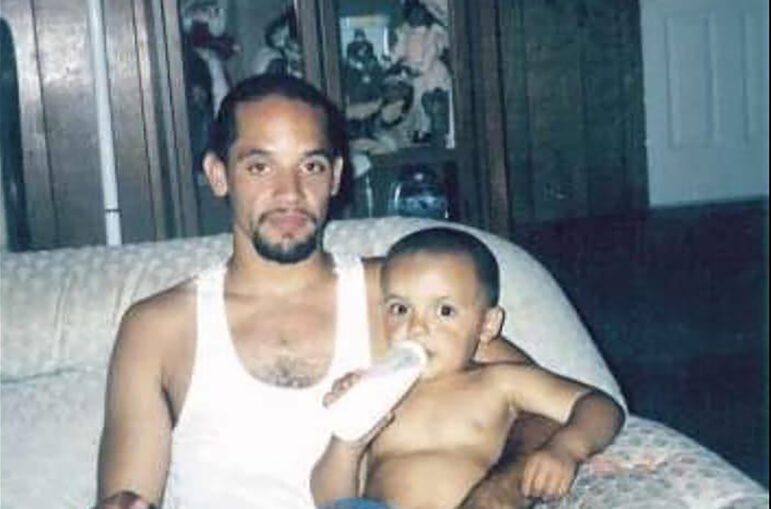 A young Black man in a white undershirt holds his baby son, who is holding a bottle of milk or formula, while sitting on a light-colored sofa.