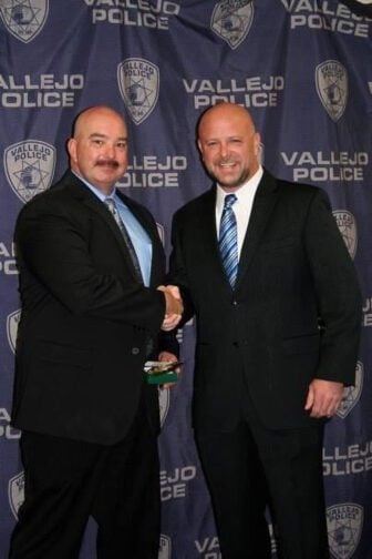 Two white Vallejo police officers with shaved heads and facial hear wearing dark suits and shaking hands.