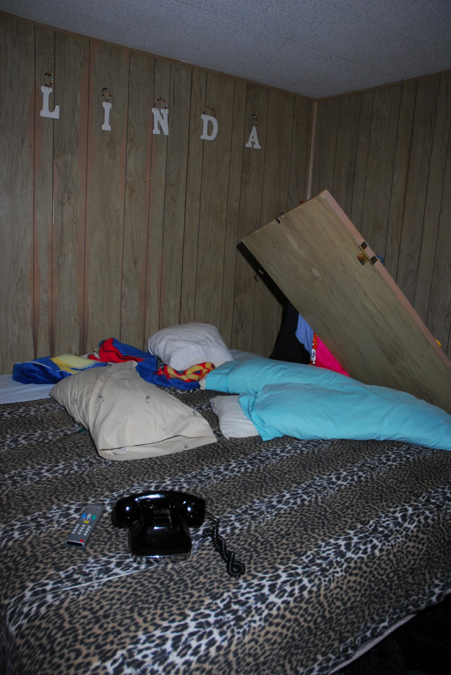 A wooden bathroom door lies against a bed covered in a faux leopard print comforter in a wood-paneled room. A black rotary phone, television remote and pillows are scattered across the bed. The name "Linda" is spelled out in plastic letters mounted to the wall.