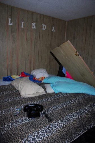 A wooden bathroom door lies against a bed covered in a faux leopard print comforter in a wood-paneled room. A black rotary phone, television remote and pillows are scattered across the bed. The name "Linda" is spelled out in plastic letters mounted to the wall.