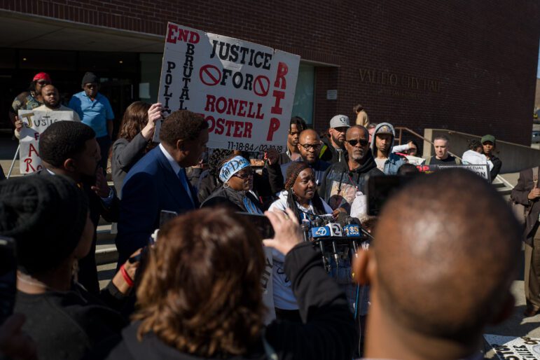 A Black woman with long braids speaks at a microphone surrounded by a diverse group of people in front of the steps to a brick city hall building outdoors during daytime. Behind her, a person is holding a large sign that reads, "Justice for Ronell Foster."