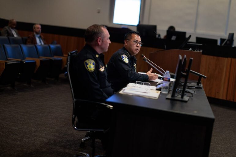 An Asian American police chief in his late 40s or early 50s speaks emphatically before a city council while seated at a black table. A captain sits to his right, listening.