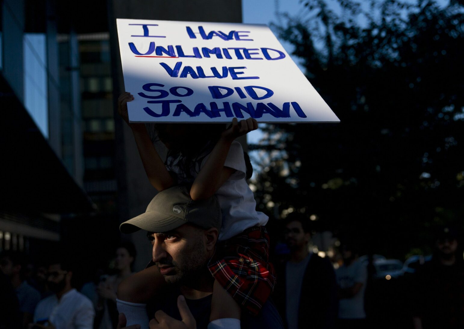 A five-year-old girl sits atop her father's shoulders holding a sign that reads, "I have unlimited value. So did Jahhnavi." They are partly in shadow and light at a protest.
