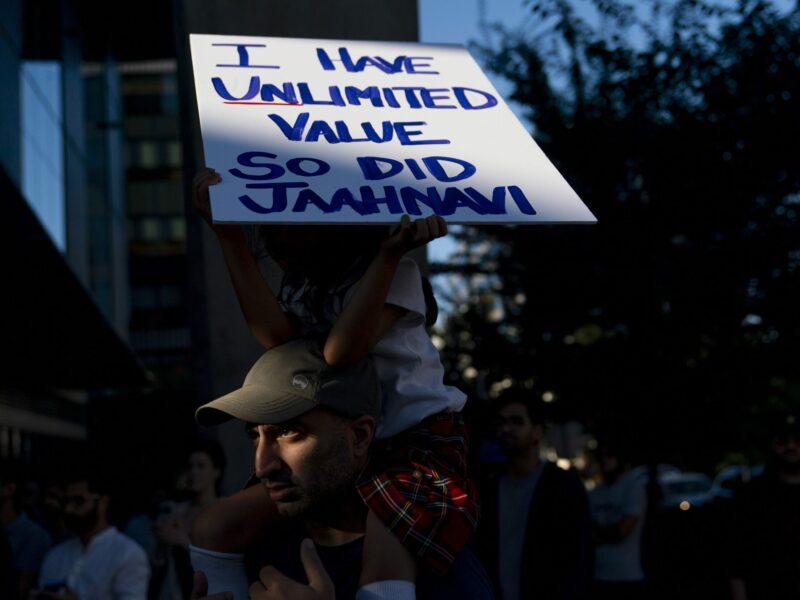 A five-year-old girl sits atop her father's shoulders holding a sign that reads, "I have unlimited value. So did Jahhnavi." They are partly in shadow and light at a protest.