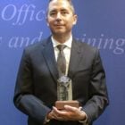 A police officer in a dark suit cradles a clear acrylic or glass award with a seven-point star and a wooden base while smiling slightly.
