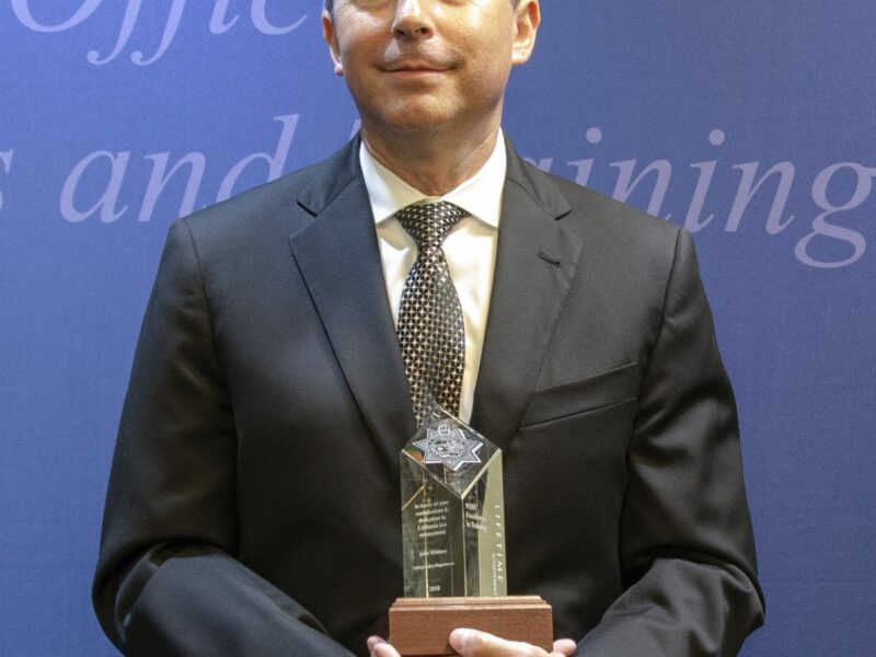 A police officer in a dark suit cradles a clear acrylic or glass award with a seven-point star and a wooden base while smiling slightly.
