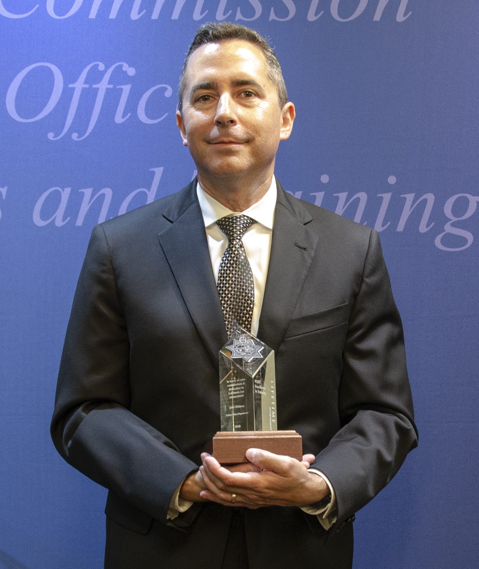 A police officer in a dark suit cradles a clear acrylic or glass award with a seven-point star and a wooden base while smiling slightly.