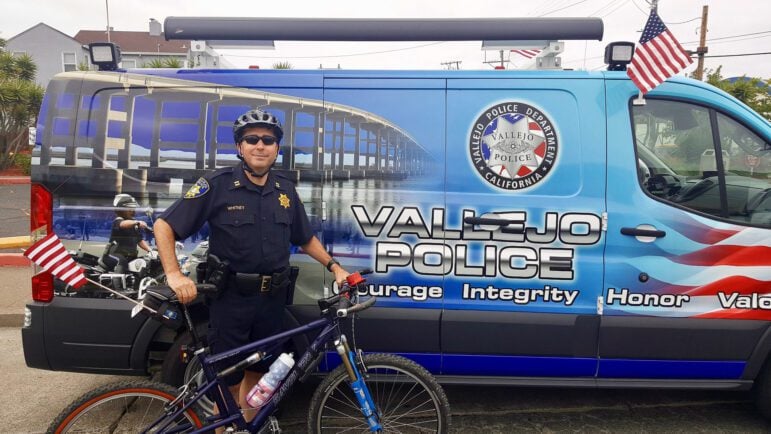 A photograph of a smiling police officer posing with a bicycle while wearing bicycle shorts and sunglasses. He is standing in front of an image-wrapped Vallejo Police Department van. Both the officer's bicycle and the van have American flags atached to it, and his bike sports an American flag water bottle.