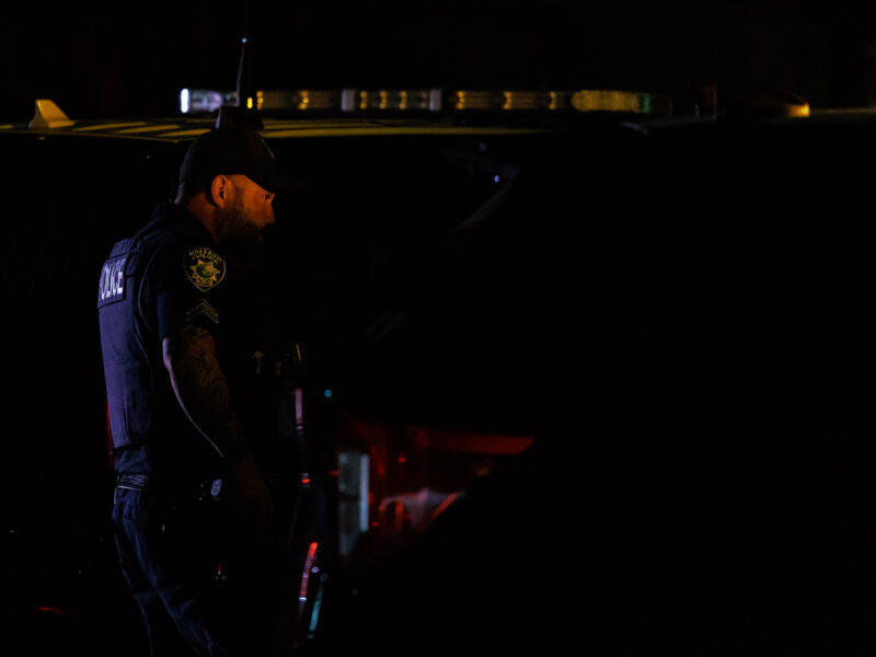 A bearded, tattooed Vallejo police officer is lit by emergency lights at night.
