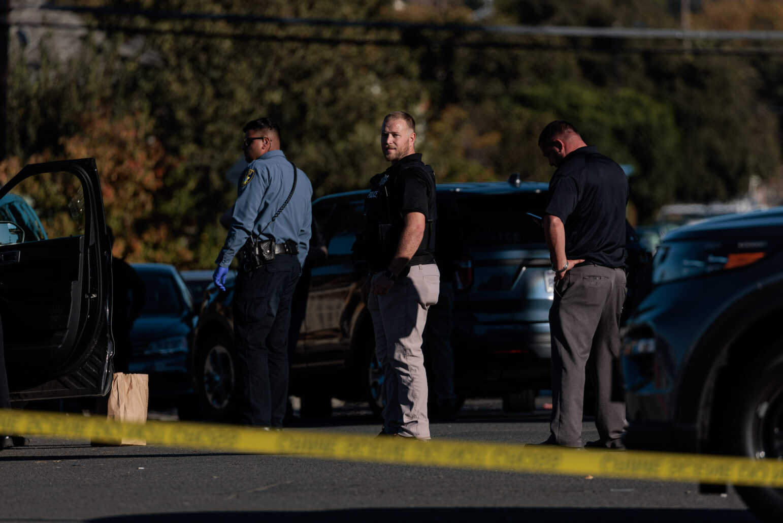Flanked by another investigator and a police assistant, a blonde police detective in khaki pants and a black shirt looks out from a crime scene during the daytime.