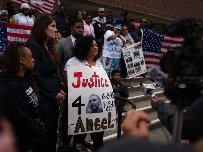 A young Latina woman with curly hair speaks to the press while surrounded by supporters on the steps of Vallejo City Hall. She holds a sign that reads, "Justice 4 Angel."