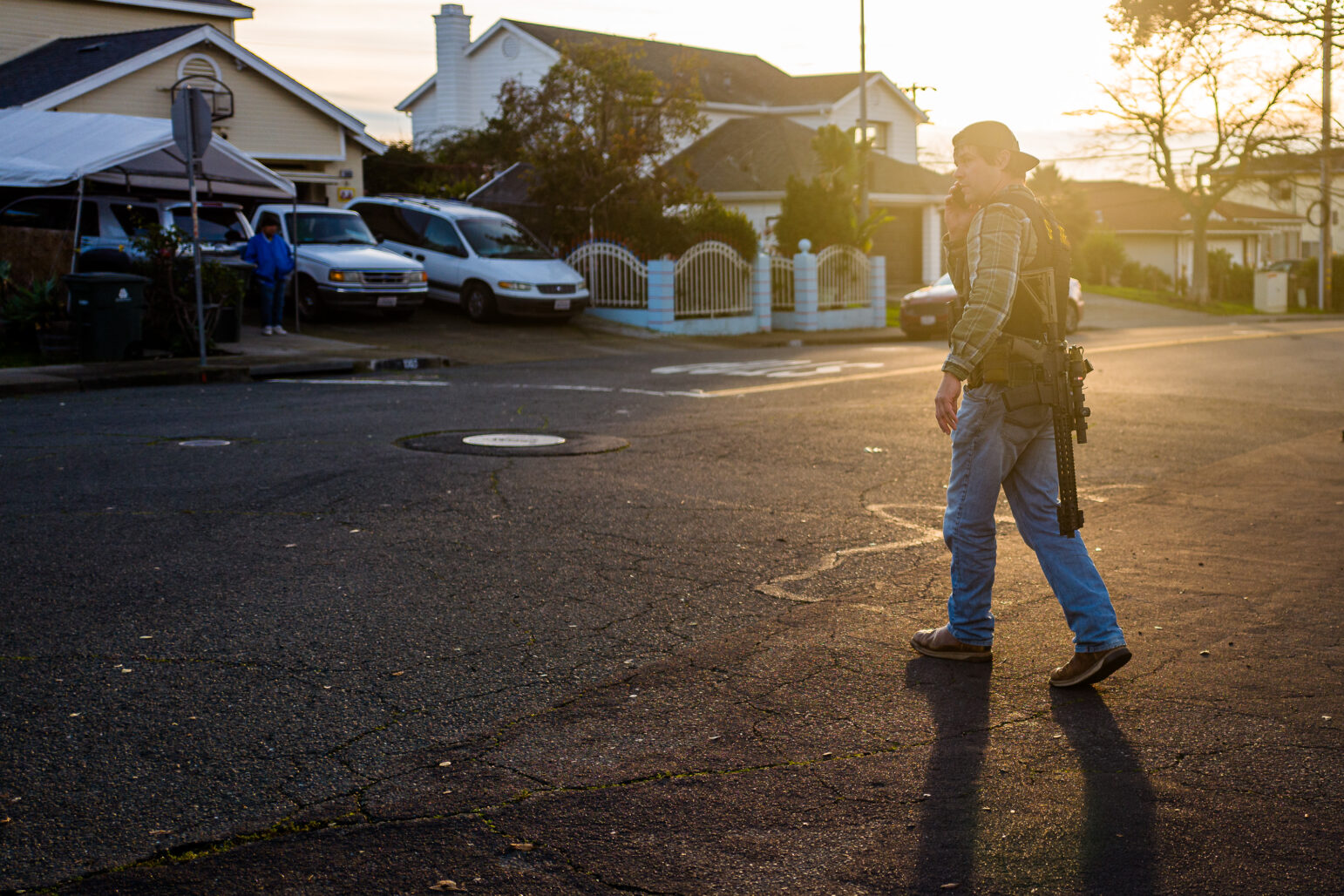 A sheriff's deputy in plainclothes with an AR-15 style rifle speaks on a cell phone at sunset.