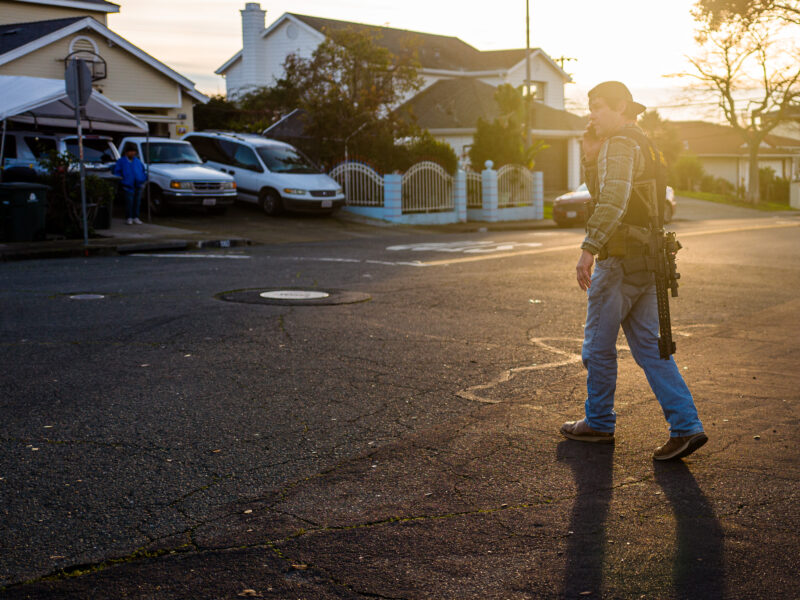 A sheriff's deputy in plainclothes with an AR-15 style rifle speaks on a cell phone at sunset.