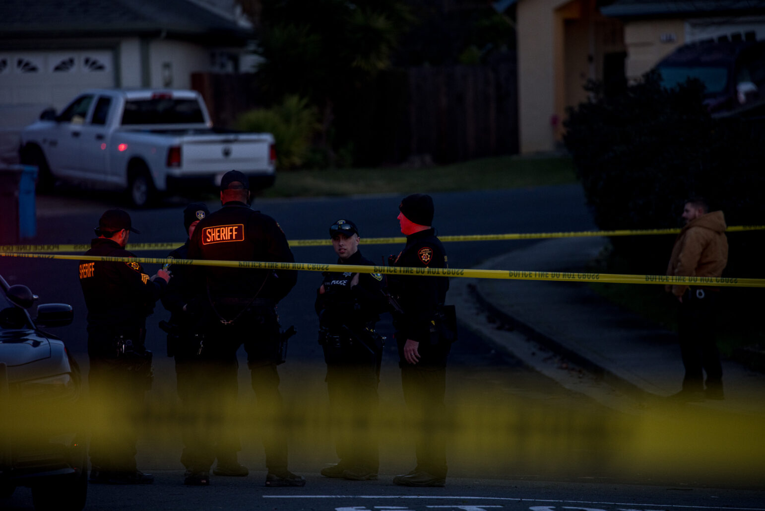A group of police and deputies stand behind crime scene tape after dusk.