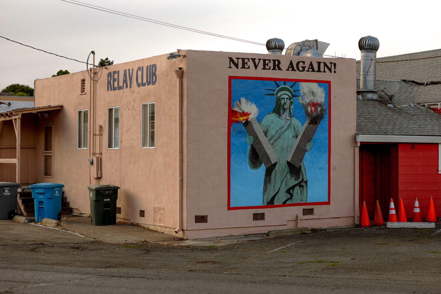 The exterior of a dive bar with the words "Relay Club" on one exterior wall and a mural of the Statue of Liberty holding the twin towers of the World Trade Center on another with the words, "Never Again."