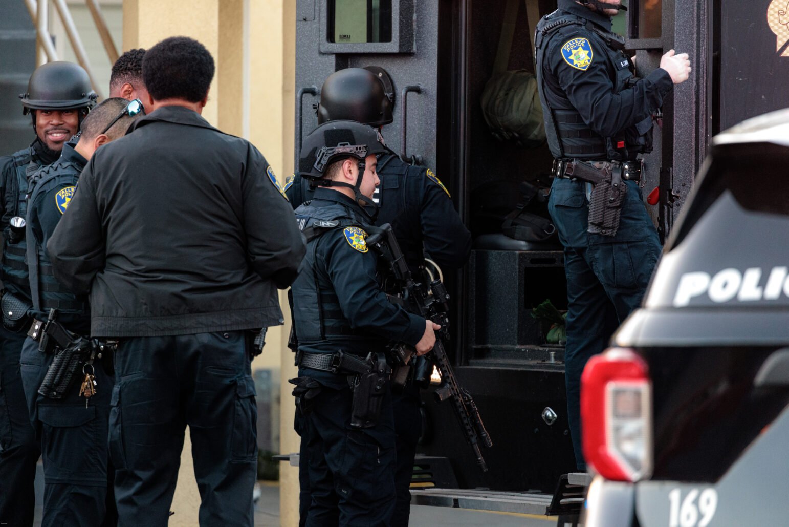 A group of law enforcement officers in tactical gear is gathered around the entrance of a vehicle, possibly preparing for a tactical operation. The officers are wearing helmets, bulletproof vests, and carrying firearms. One officer is looking towards the camera and smiling, amidst a serious and focused setting. This officer is identified as Ofc. Ronald Dupree from the Vallejo Police Department. There is a marked police vehicle in the foreground.