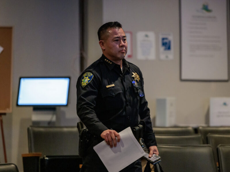 A uniformed police chief walks through a city council chambers with rows of empty chairs, holding papers in one hand and a smartphone in the other. He wears a dark uniform with a badge, name tag, and radio attached to his shoulder. His duty belt is equipped with various gear, including a holstered sidearm. The background includes informational posters on the wall.