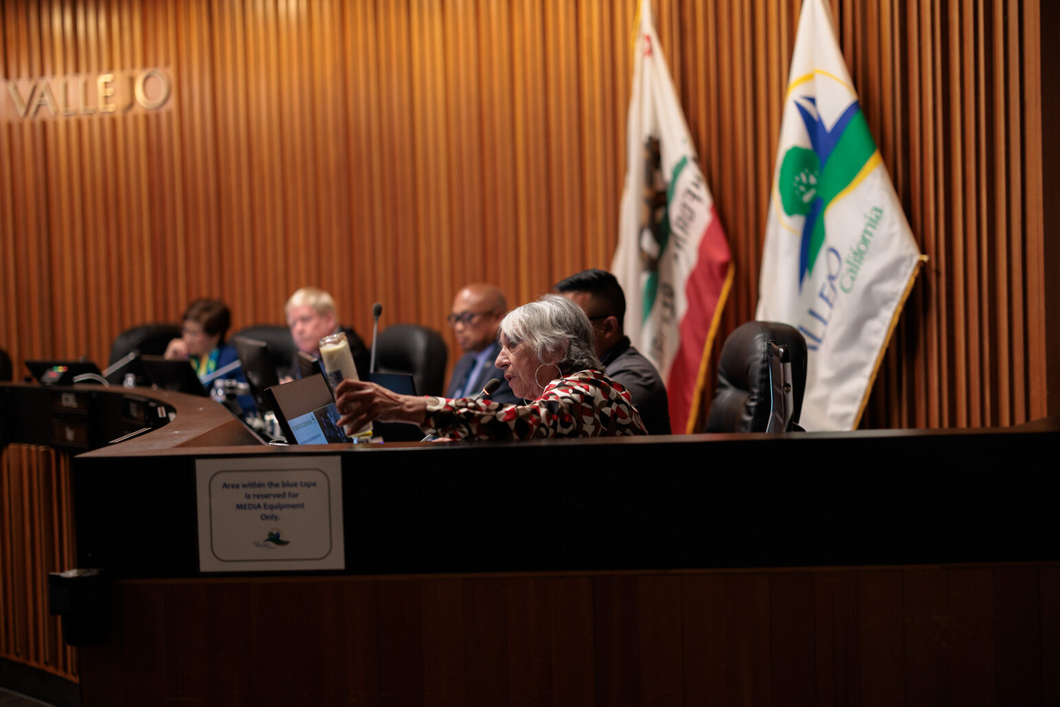 A Vallejo city councilwoman with white hair and wearing a brightly patterned top places a votive candle on the dais in front of her.