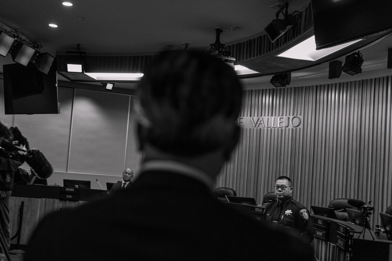 A black and white photo taken from behind a person's head, overlooking a city council chamber where a uniformed police officer is seated at the dais. The room is equipped with professional lighting and video recording equipment, indicating a formal proceeding or event. The words 'CITY OF VALLEJO' are prominently displayed on the wall behind the dais. The focus is on the officer and the surroundings, with the foreground figure's head slightly blurred.