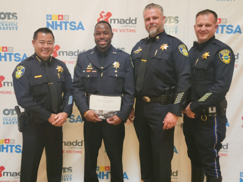 Four police officers are standing together, smiling in front of a backdrop with various logos, including those of OTS (California Office of Traffic Safety), NHTSA (National Highway Traffic Safety Administration), and MADD (Mothers Against Drunk Driving). One officer is holding a certificate, which he earned for making 19 impaired driving arrests in the previous year.
