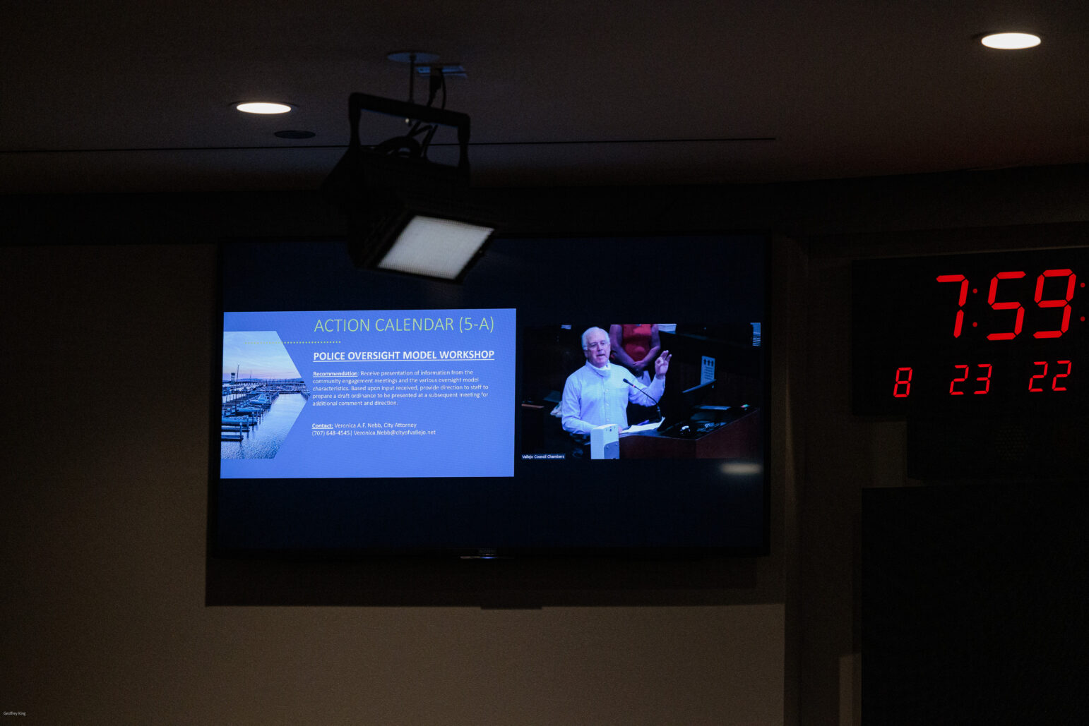 An image showing a darkened room with a large digital display screen, featuring a slide from a presentation titled 'Police Oversight Model Workshop'. On the screen, a man is pictured speaking at a podium. To the right, there's a digital clock displaying the time as 7:59 and the date as August 23, 2022, in red numerals, indicating the context of a formal meeting or conference setting.