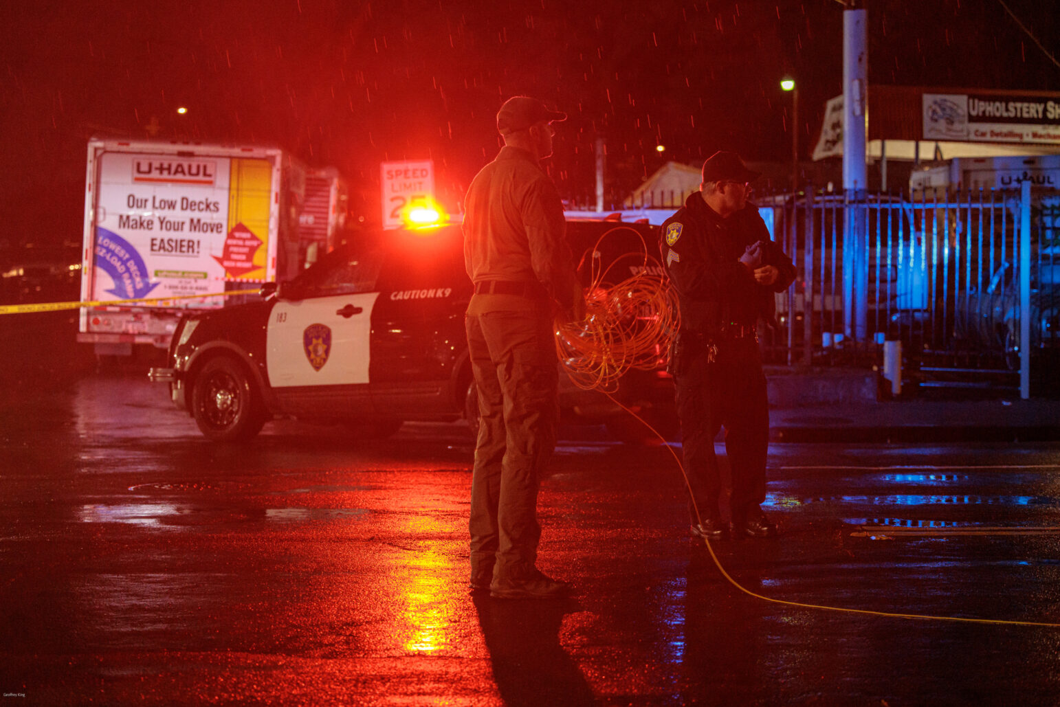 A night-time scene capturing two men on a rain-soaked street, illuminated by the red and blue lights of a police vehicle. A sheriff's deputy is holding what appears to be a coil of detonation cord, while the another man, a Vallejo police officer, observes the scene. The wet pavement reflects the colorful lights, adding to the dramatic atmosphere.