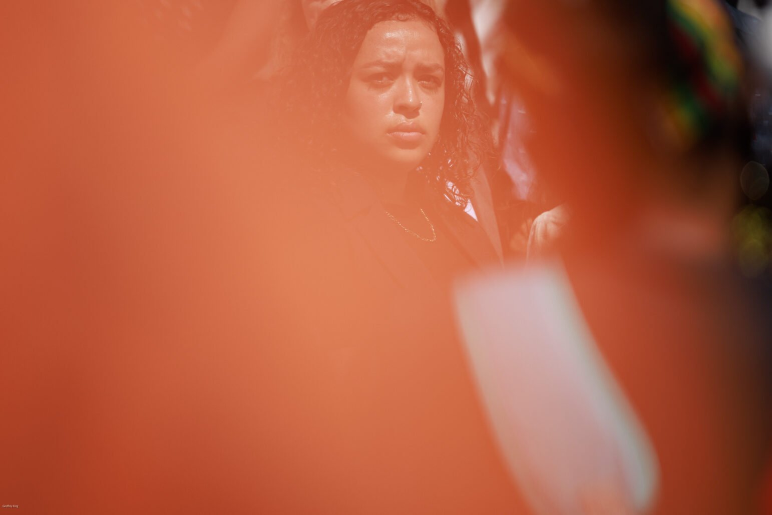 A woman is partially obscured by a bright orange, translucent foreground, creating a dreamlike effect. She appears contemplative and is dressed in professional attire, with her curly hair framing her face, amid a crowd at an outdoor event.